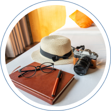 A straw hat with a black ribbon sits on a white bed next to a brown leather notebook with a pen, a pair of black glasses, and a vintage camera with a leather strap. An orange pillow and curtains are visible in the background.